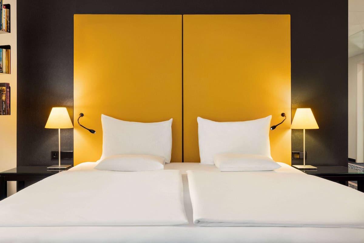 A double bed with white linens, two bedside lamps, and a large, yellow accent wall.
