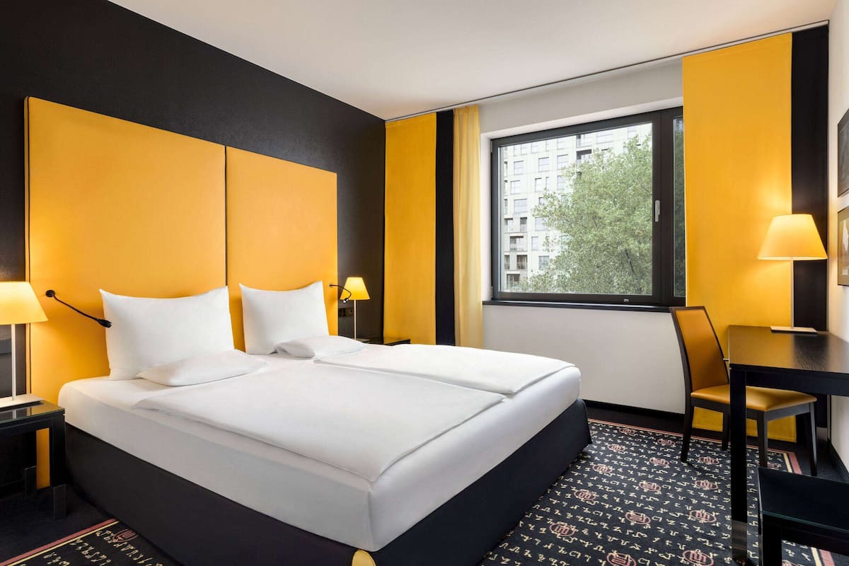 A hotel room with a large bed, two bedside tables with lamps, a desk with a chair, and a window with a view of buildings and trees.