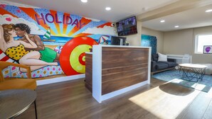 Un espace d’accueil agrémenté d’une fresque murale colorée, d’un comptoir de réception en bois, d’un canapé gris et d’un téléviseur fixé au mur.
