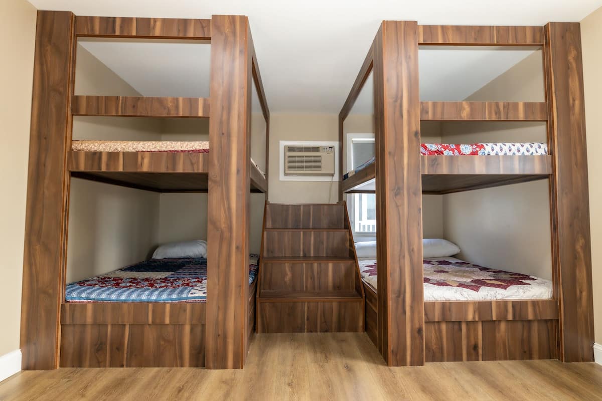 Una habitación con tres literas de madera, una escalera en el centro y un pequeño ventanal con aire acondicionado.