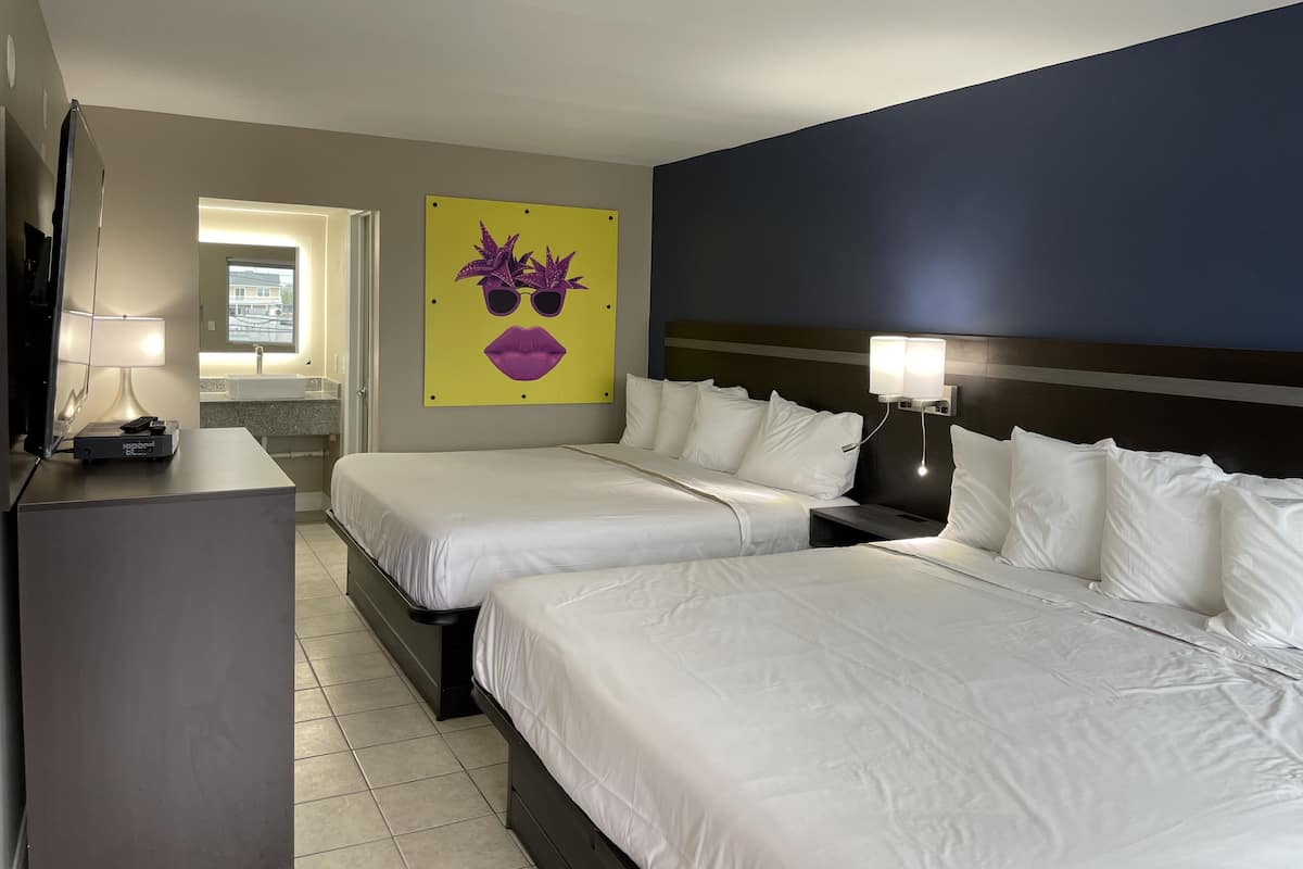 Habitación de hotel con dos camas, un escritorio, una lámpara y un baño visible a través de una puerta abierta.