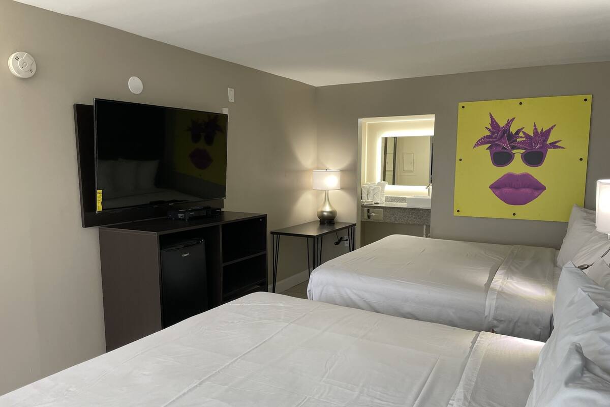 Habitación de hotel con dos camas, televisión, escritorio, lámpara, espejo y baño con lavabo y espejo.