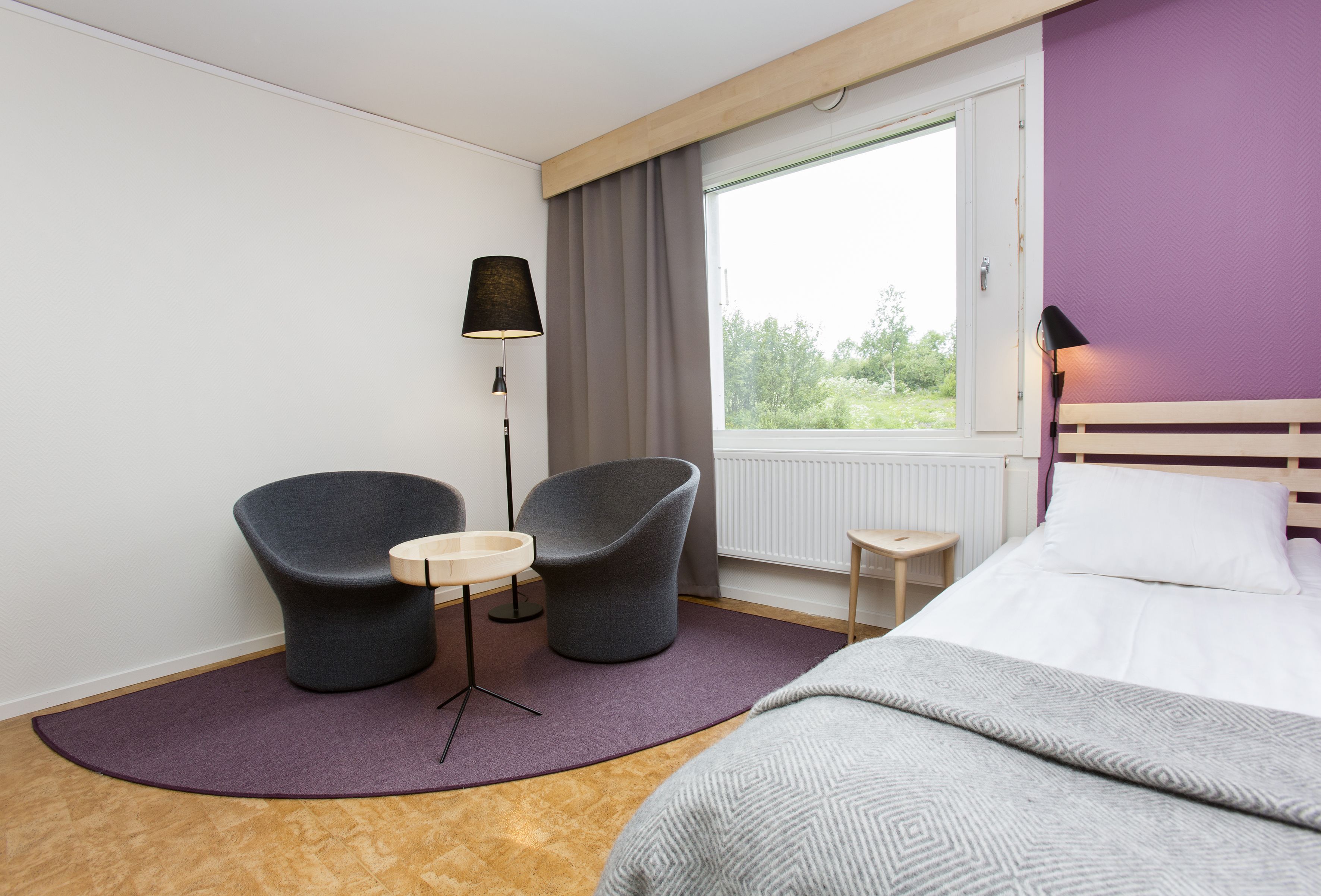 A hotel room with a bed, two armchairs, a small table, a lamp, and a window with a view of greenery.
