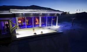 Area piscina esterna innevata con un recinto in vetro, una vasca idromassaggio e una zona relax con vista sulle montagne.