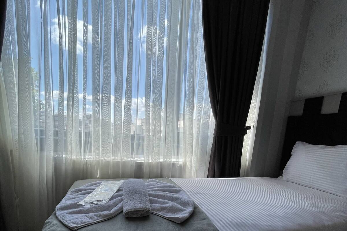A hotel room with a bed, a window with sheer curtains, and a view of the sky.