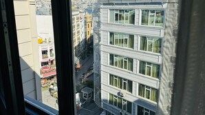 Vista desde una ventana que da a un edificio de varios pisos con balcón, una calle abajo y cielo despejado.