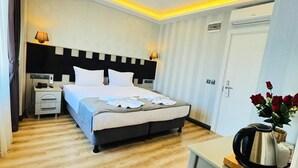 Una habitación de hotel con cama, mesitas de noche, un escritorio con hervidor y un jarrón con flores.
