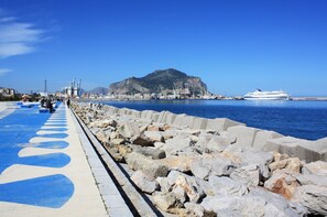 A coastal promenade with a rocky breakwater, a clear blue sky, and a cruise ship in the distance.