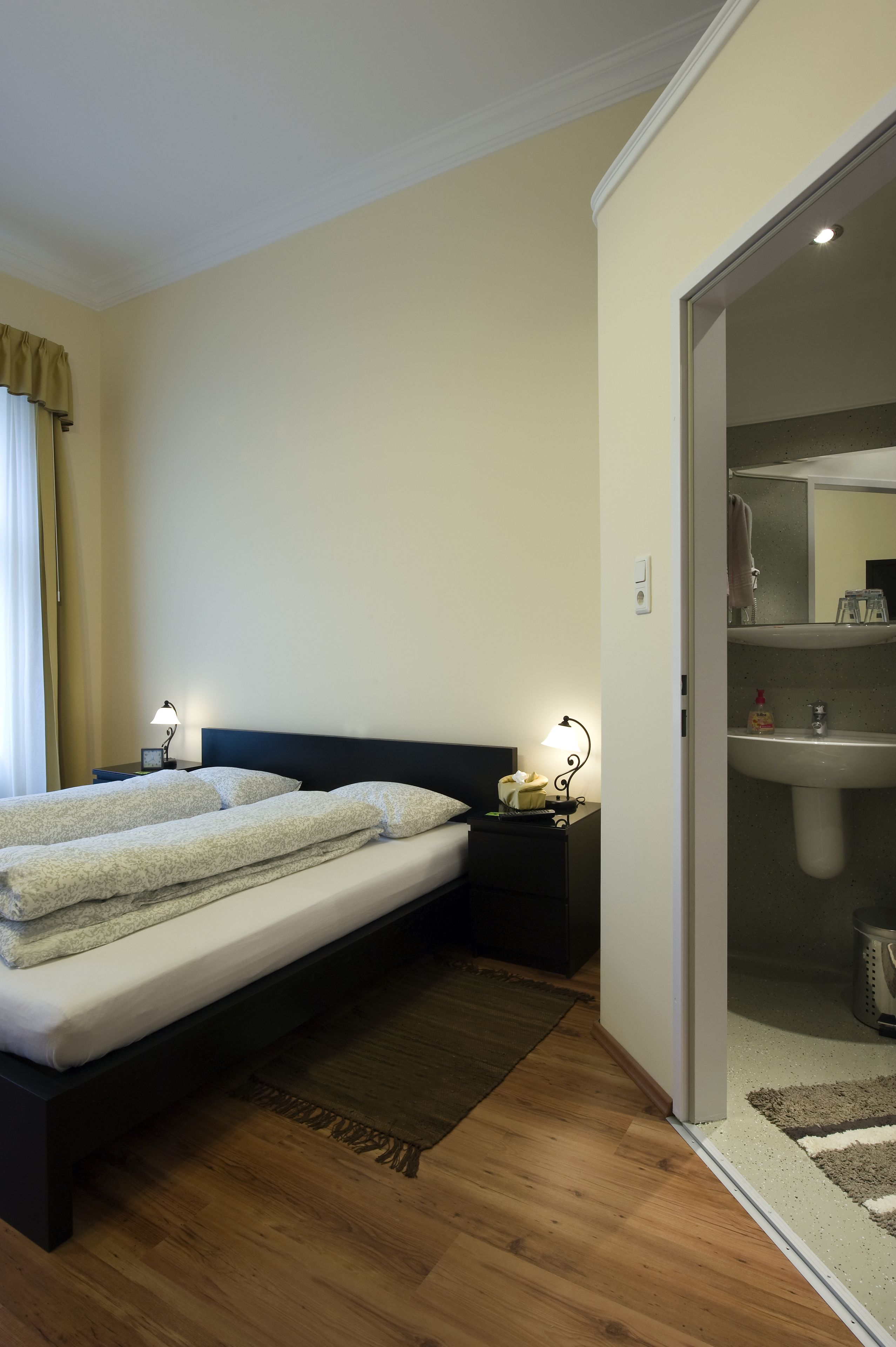 Habitación de hotel con cama doble, lámparas de noche, mesita de noche, suelo de madera y baño con lavamanos y espejo.