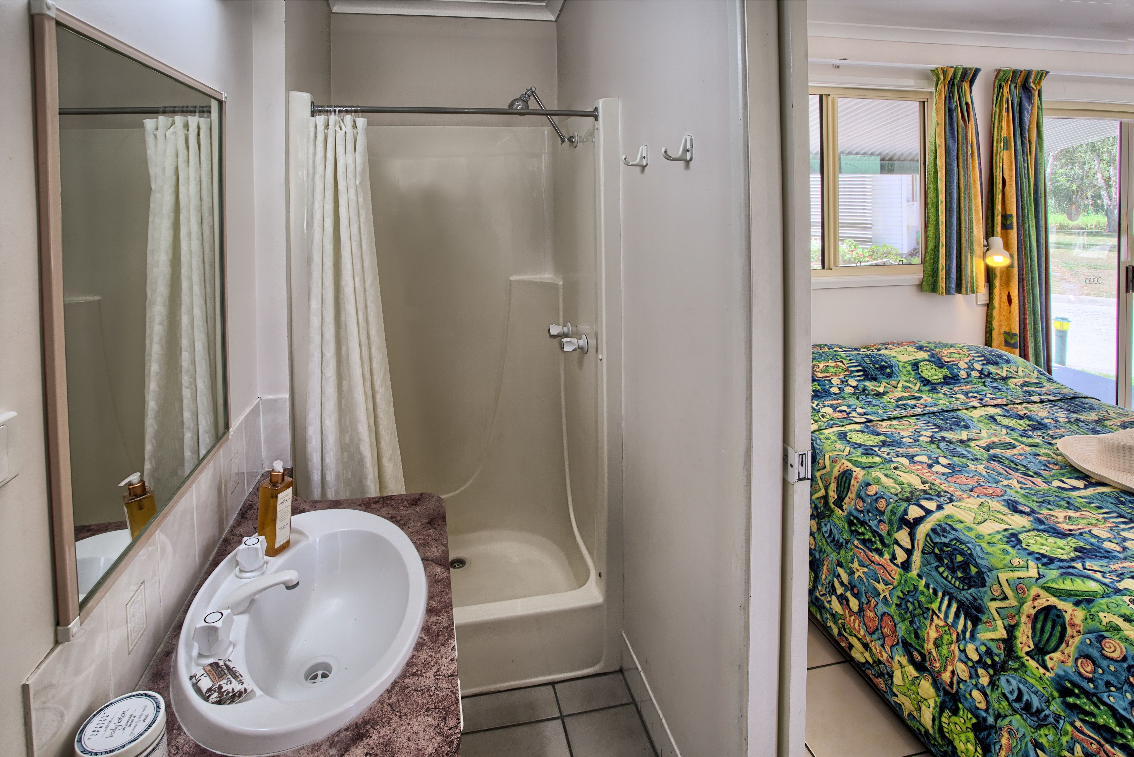 A bathroom with a sink, mirror, shower, and a bed with a colorful bedspread.