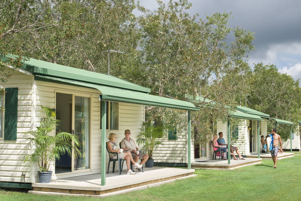 A row of small, single-story cabins with green roofs and open porches, surrounded by trees.