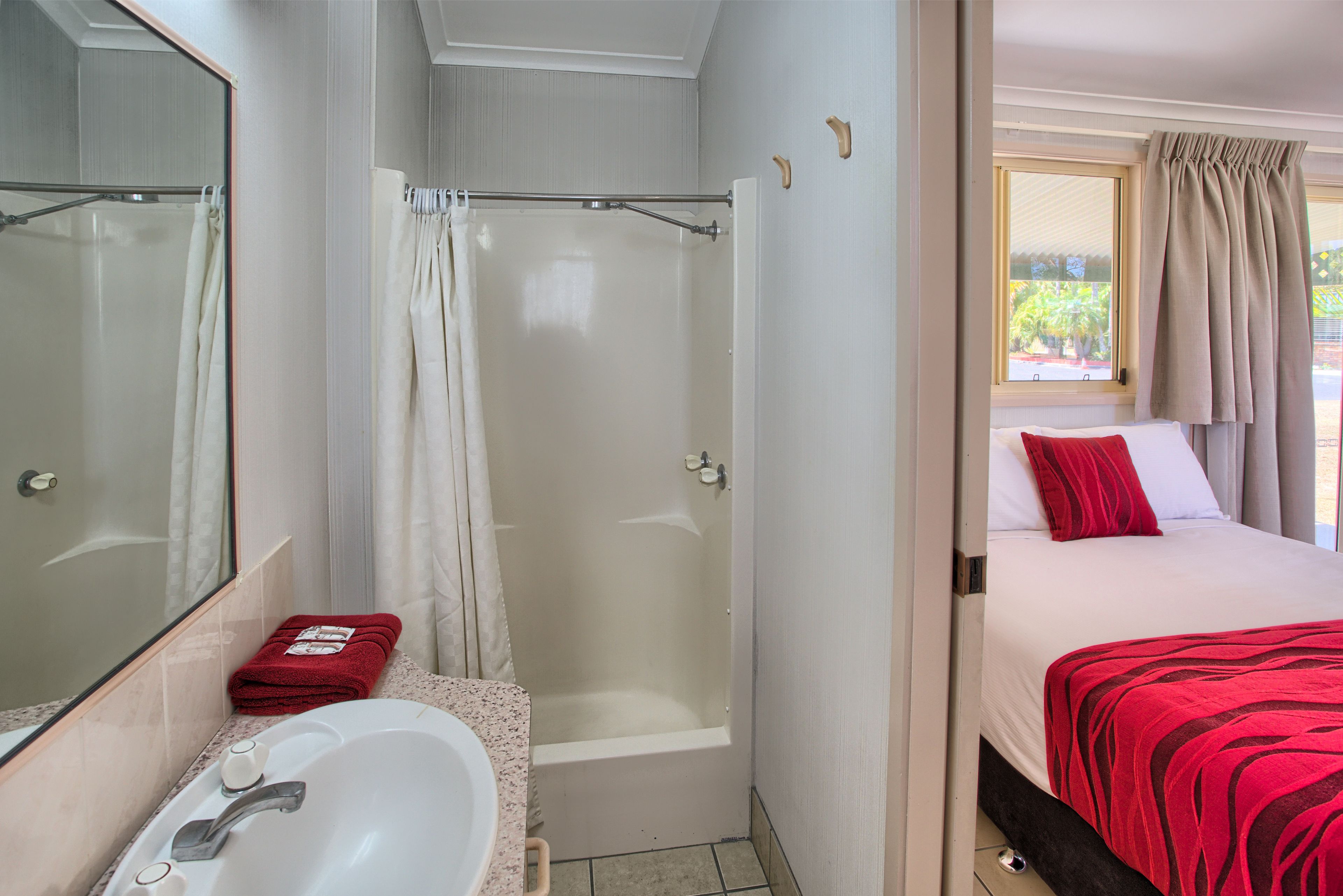 A bathroom with a bathtub, a shower, and a sink. There is a bed with red bedding and a window with curtains.