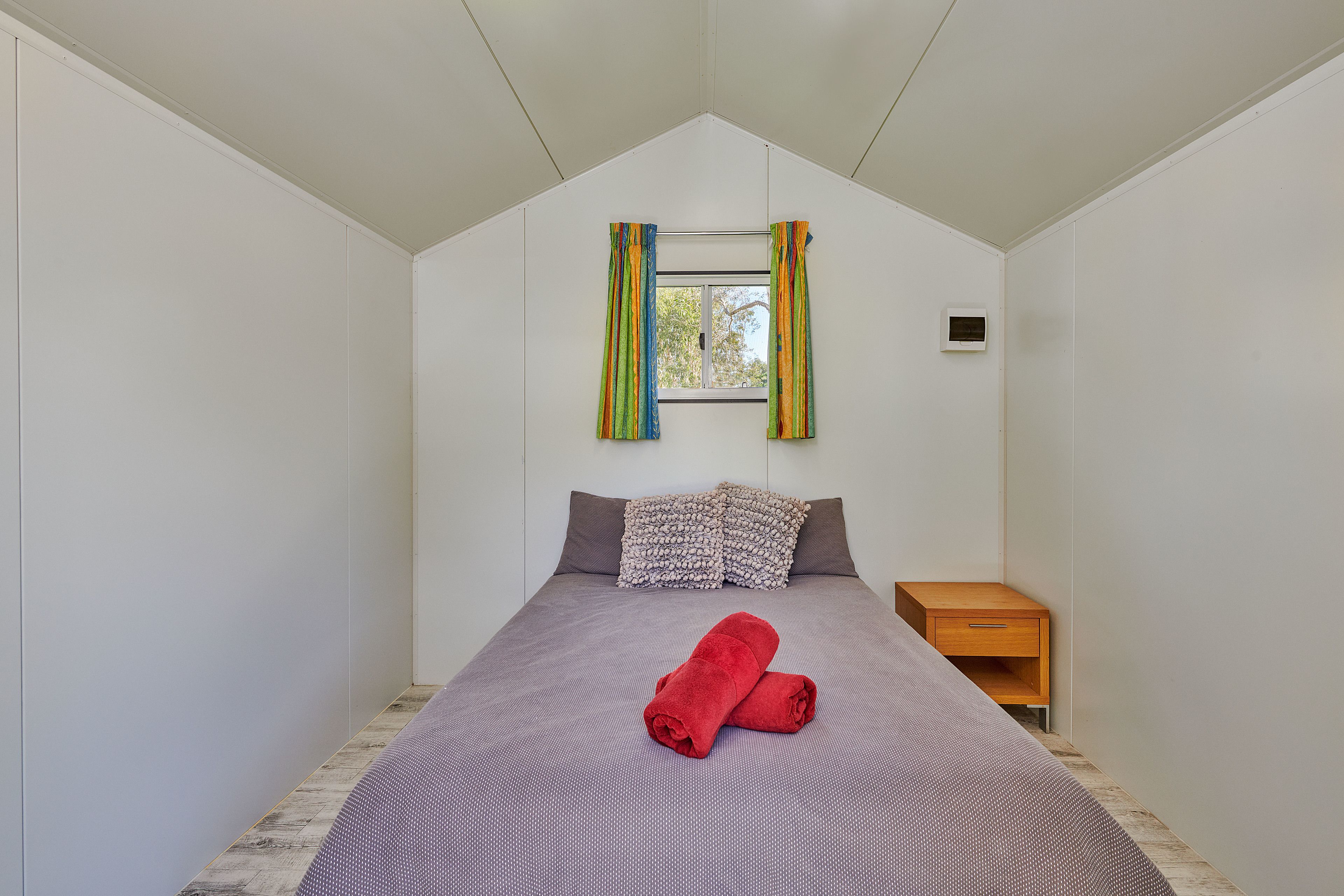 A bedroom with a bed, a window with curtains, a bedside table, and a wall-mounted thermostat.