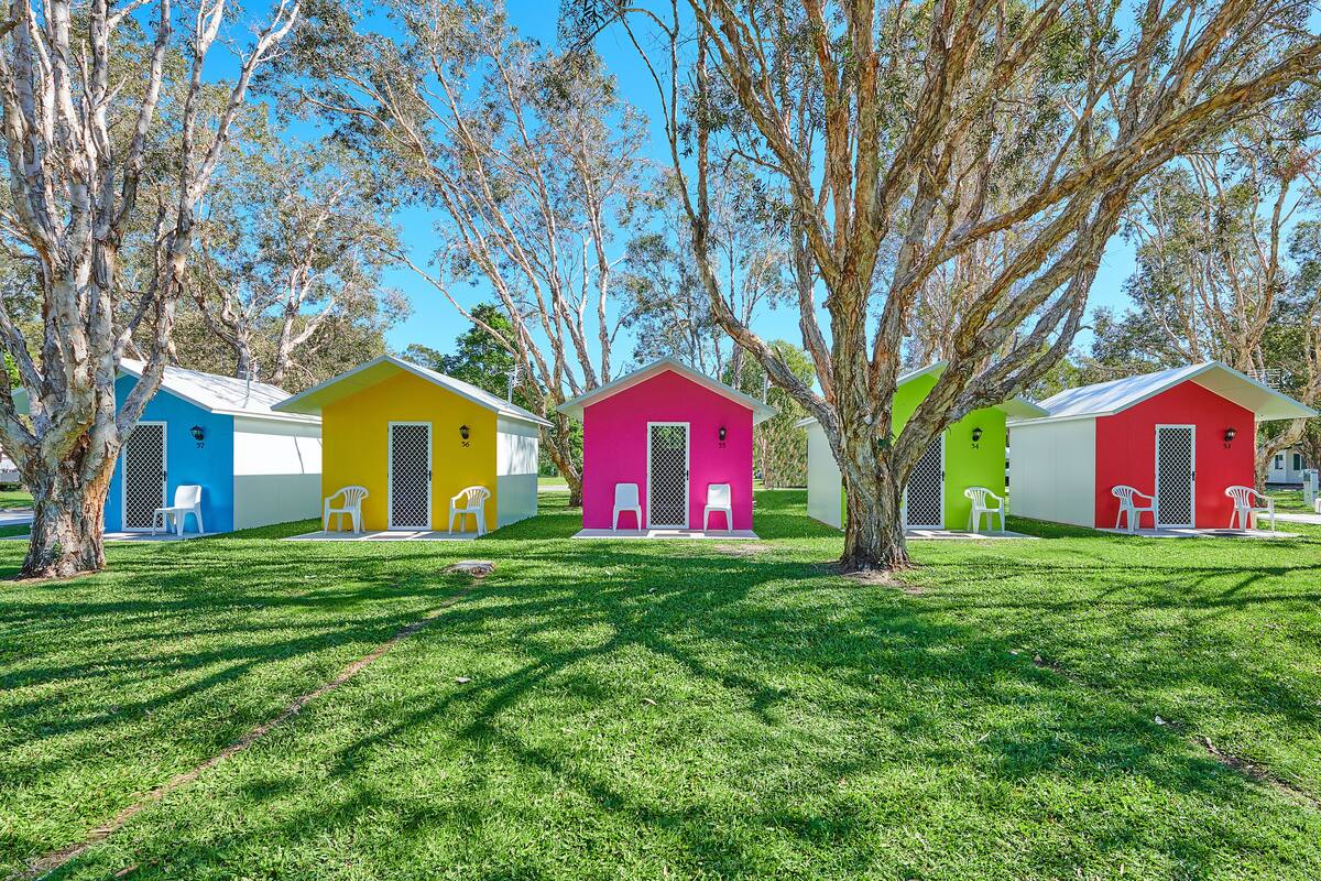 A row of colorful cabins with white chairs outside, surrounded by trees.