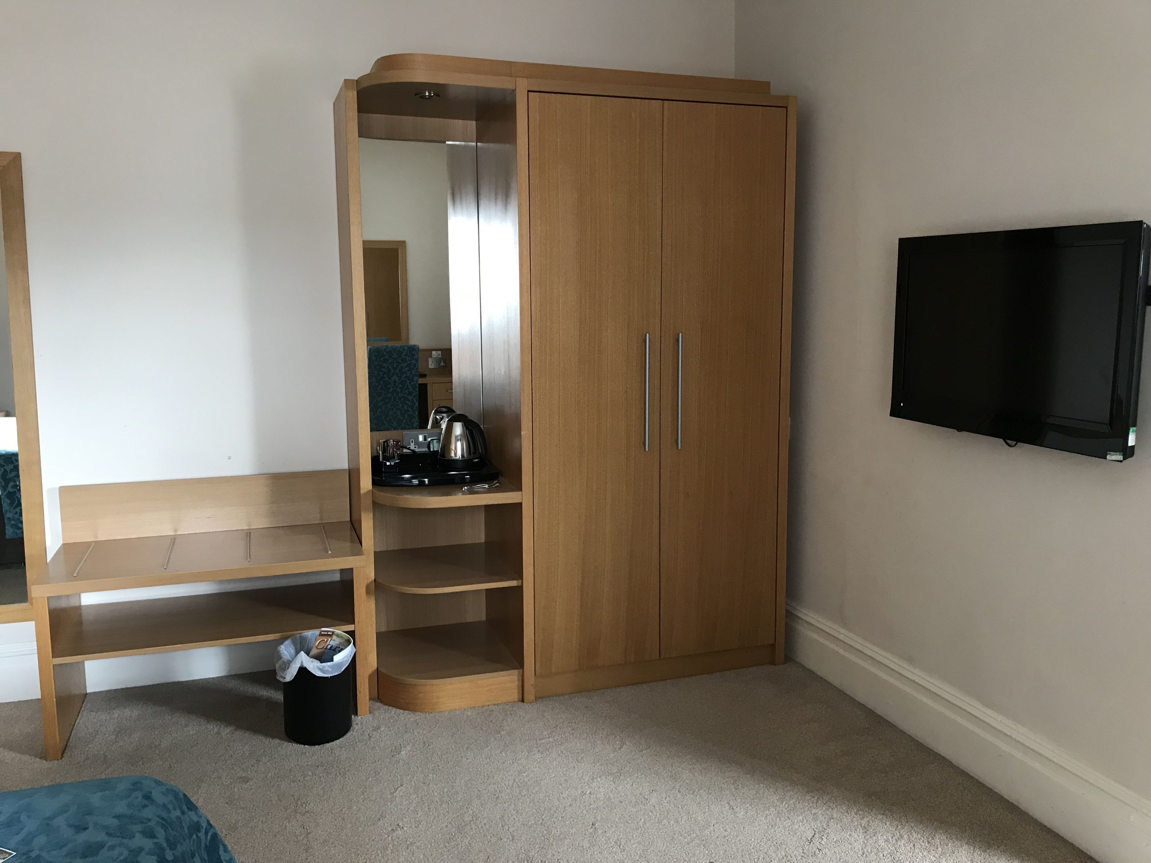 A room with a wooden wardrobe, a mirror, a television, a desk, and a trash bin.