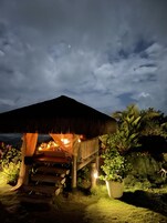 A thatched-roof structure with a wooden staircase leading up to it, surrounded by lush greenery and a large potted plant.