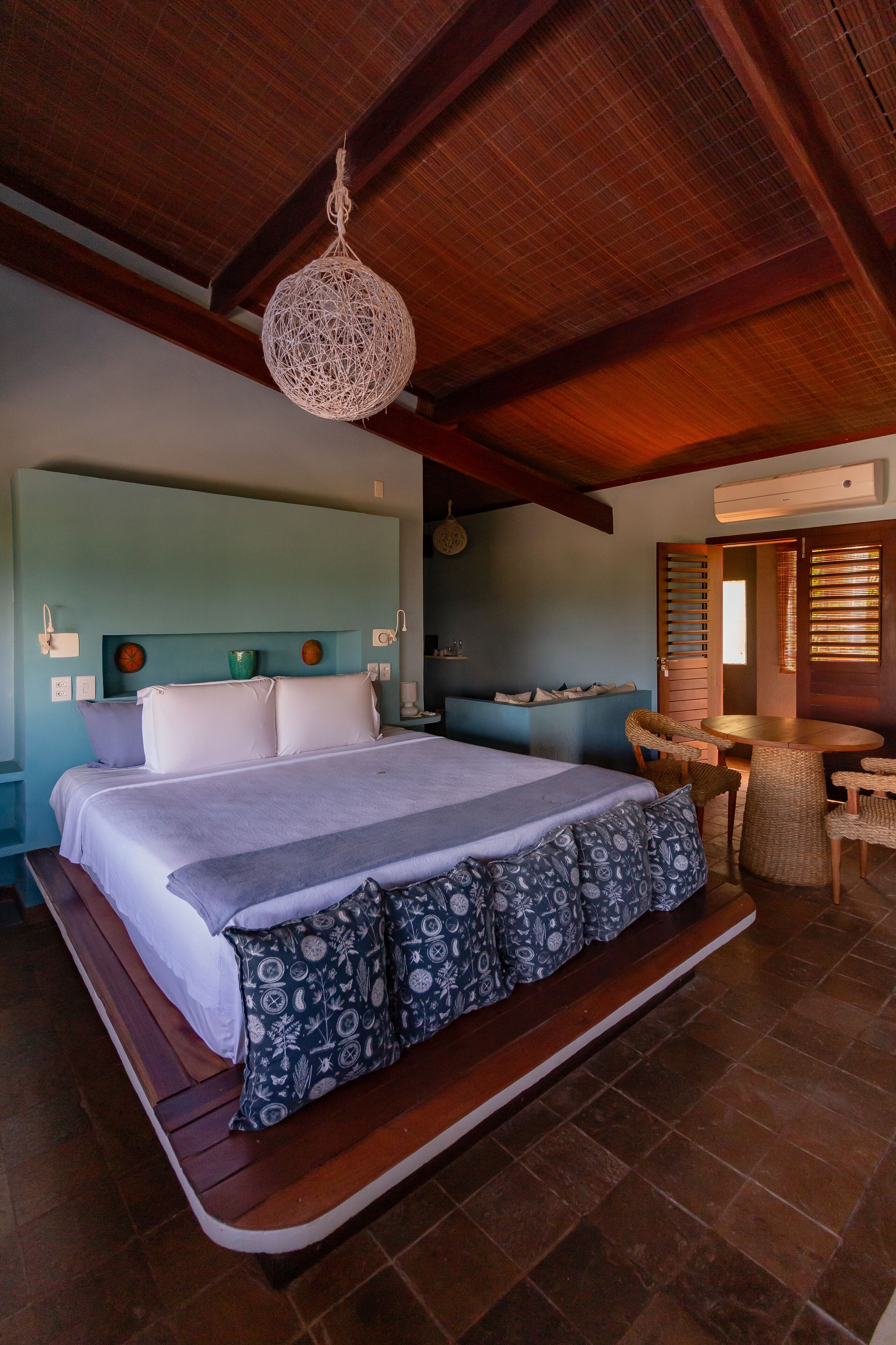 A spacious bedroom with a large bed, a hanging light fixture, a small seating area, and a bathroom visible through an open door.