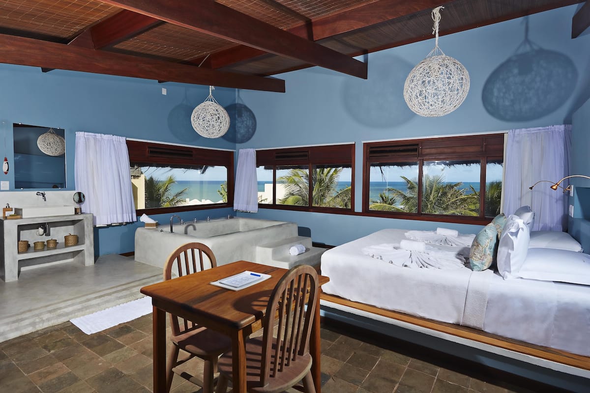 Una habitación con cama, mesa, sillas, lavamanos y vista al océano.