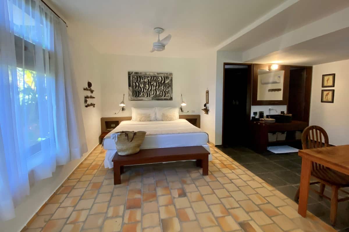 Un dormitorio con cama, mesitas de noche, un banco, ventilador de techo, una obra de arte en la pared y un baño visible a través de una puerta abierta.