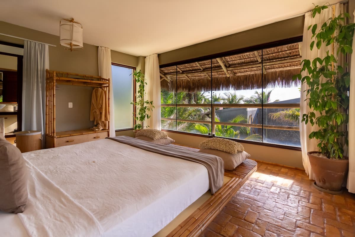 Un dormitorio espacioso con una cama grande, un cabecero de madera, una ventana con vistas a un tejado de paja y una planta en maceta.