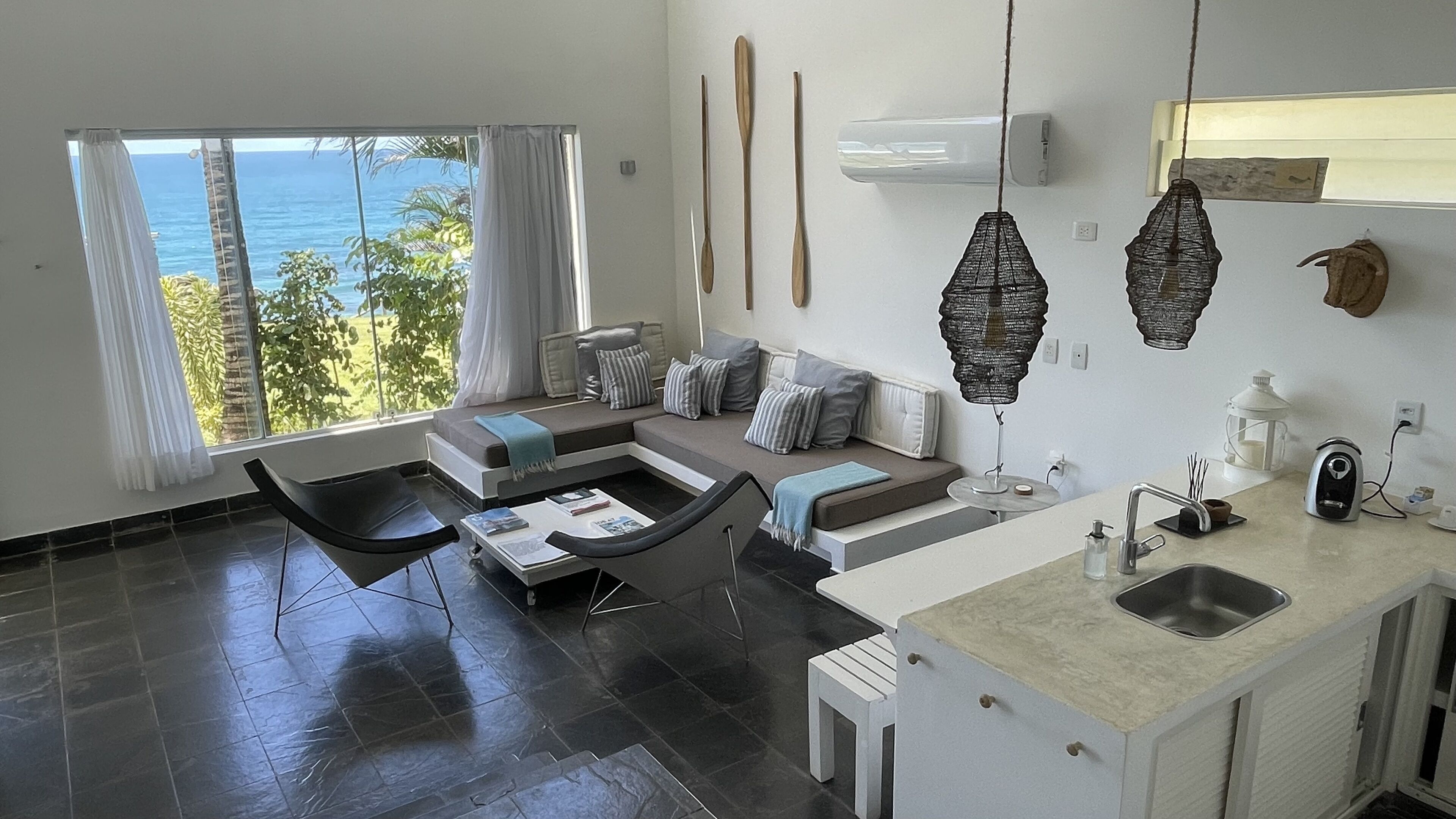 A modern living room with a large window, a sofa set, a coffee table, and a kitchen area with a sink and a kettle.