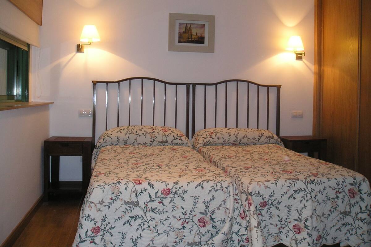 Una cama doble con ropa de cama floral, dos mesitas de noche, una mesita auxiliar y un cuadro enmarcado en la pared, además de una puerta de madera.