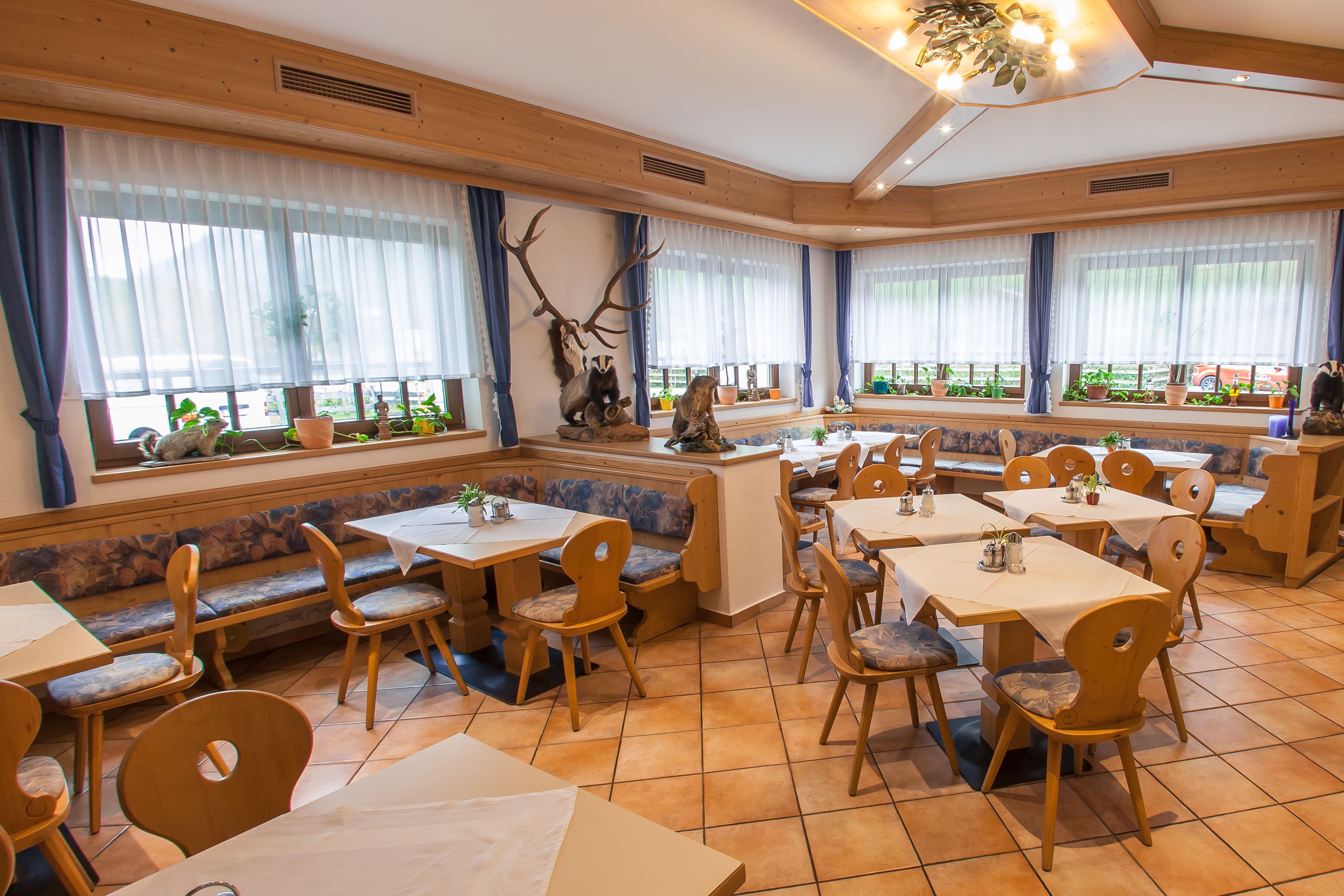 Un restaurante con mobiliario de madera, amplias ventanas con cortinas y una variedad de plantas.