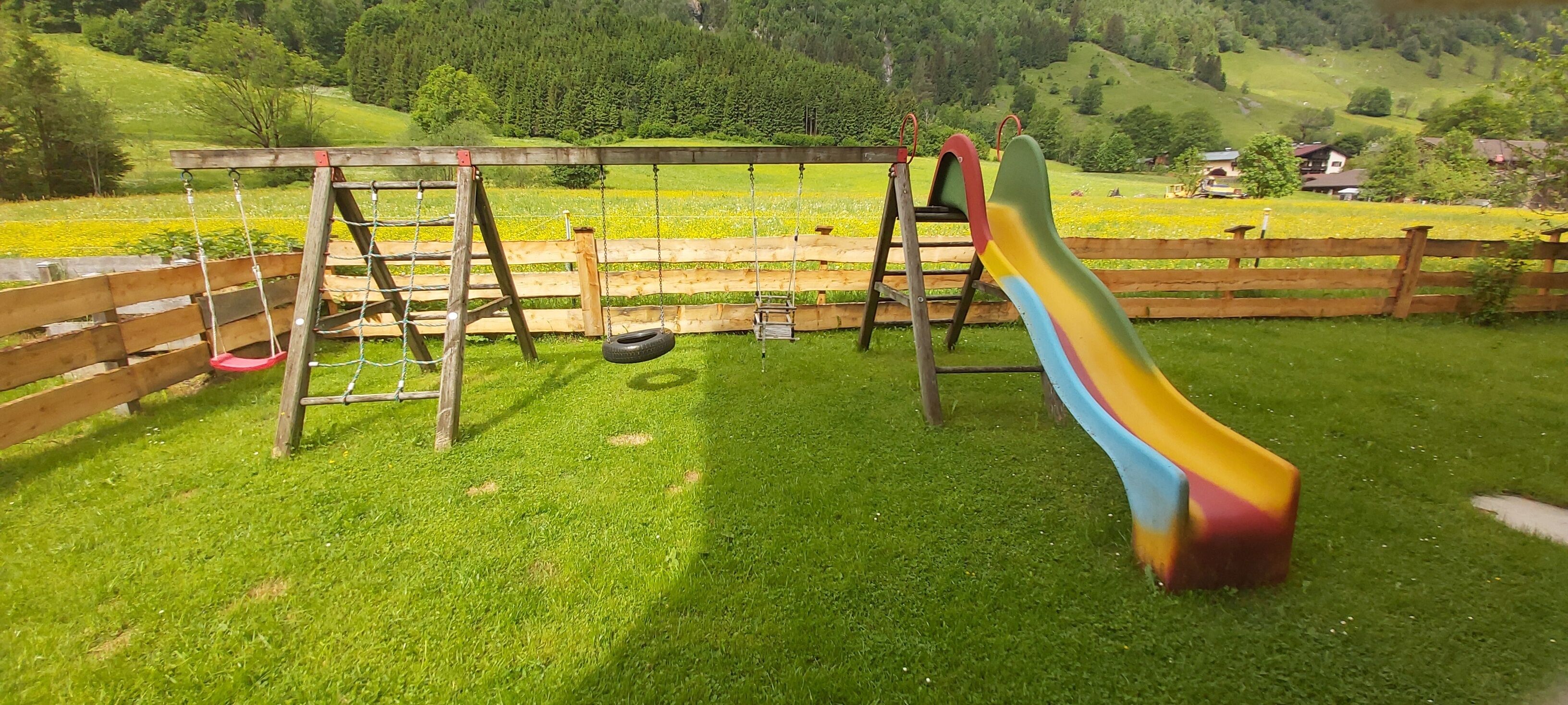 Un parque infantil con columpios y un tobogán en un área verde con cerca de madera.