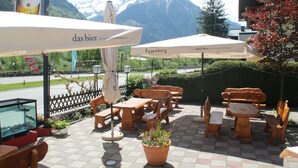 An outdoor seating area with wooden tables and chairs, umbrellas, and a fish tank. - Hotel Wasserfall (Fusch an der Grossglocknerstrasse)