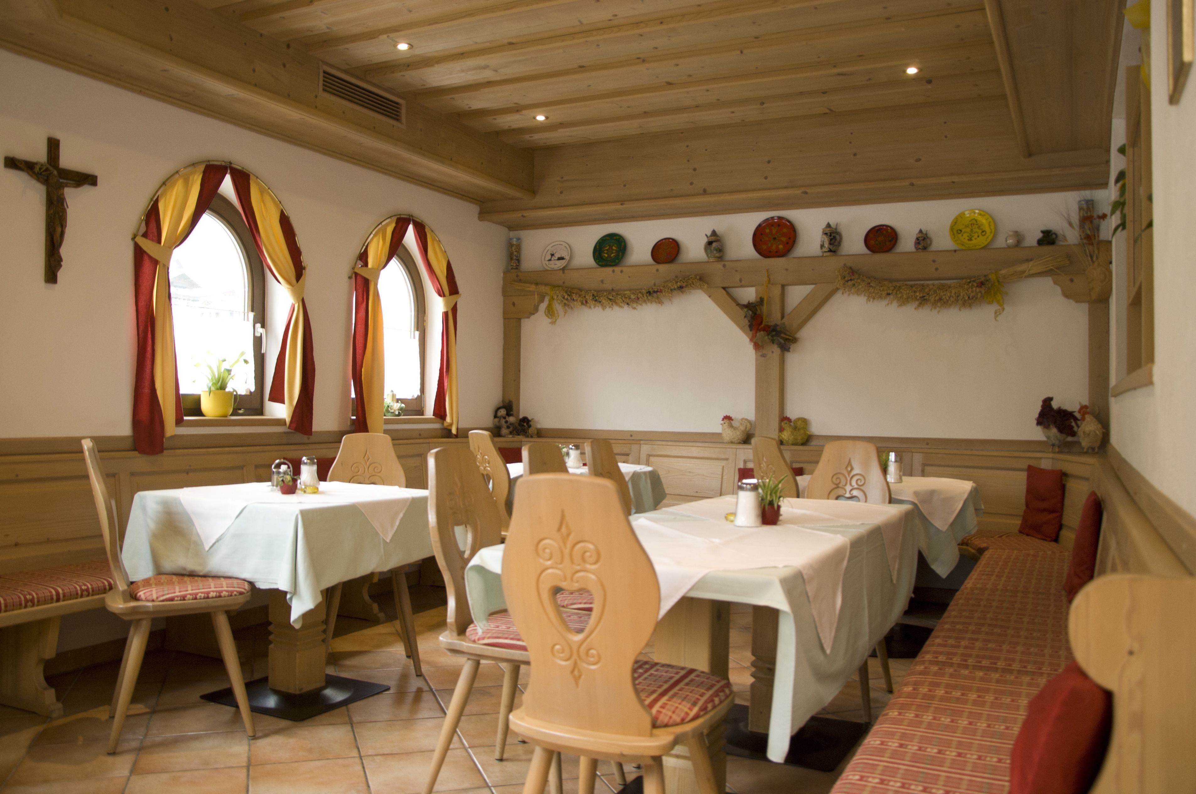 Un comedor con mesas y sillas de madera, una cruz en la pared y una repisa con platos y adornos.