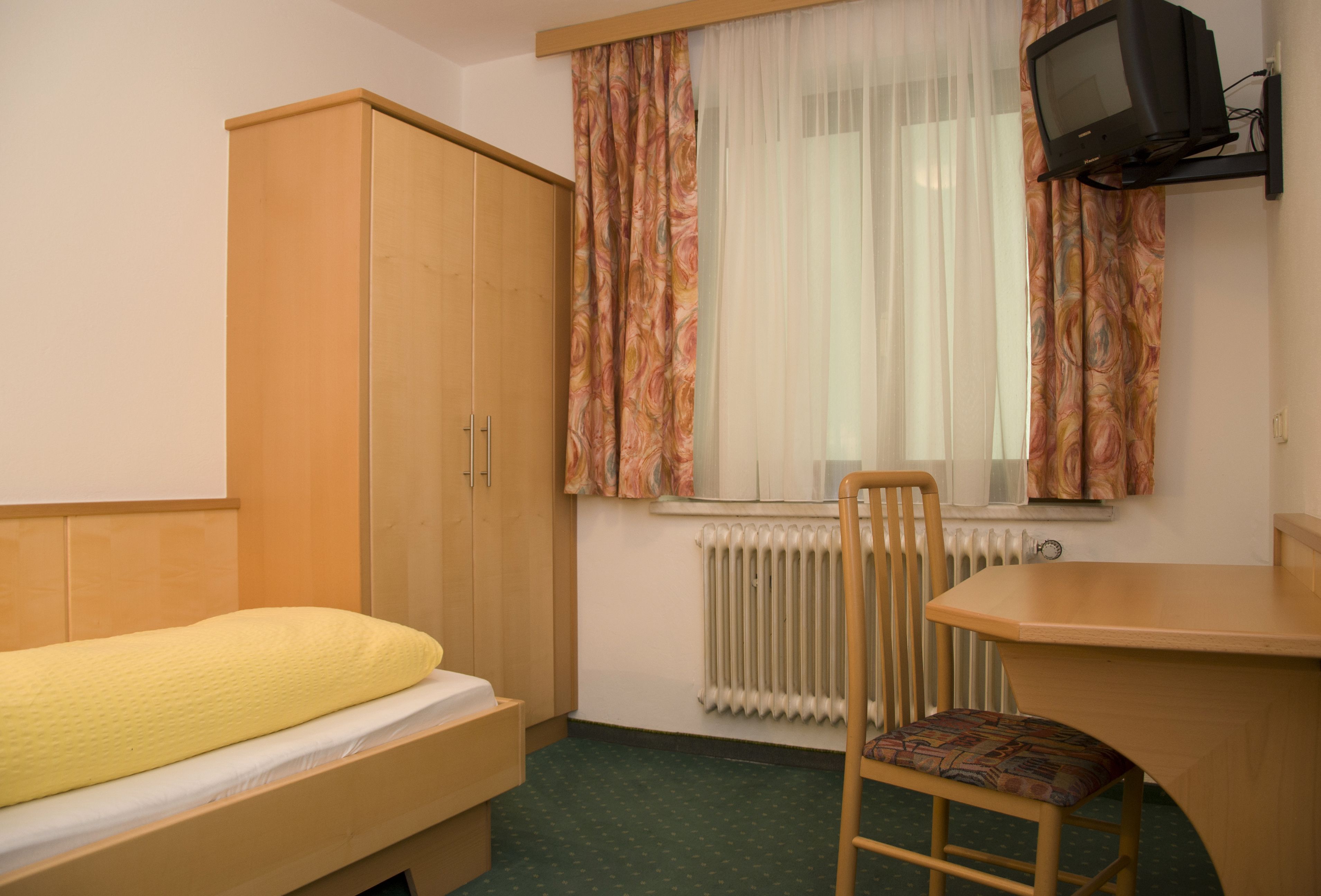Une petite chambre d’hôtel composée d’une seule pièce, comprenant un lit, une armoire en bois, un bureau avec une chaise, un téléviseur et une fenêtre avec des rideaux.