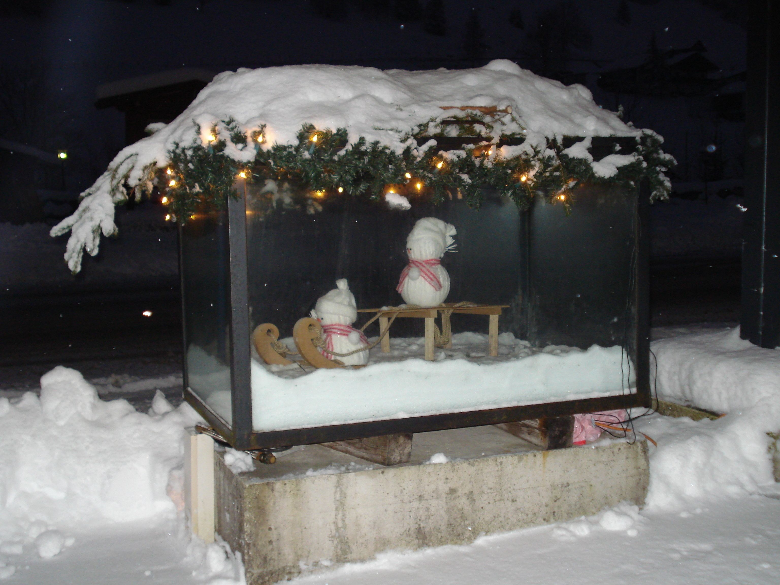 Una vitrina de cristal cubierta de nieve con un muñeco de nieve y un reno, decorada con luces y follaje.