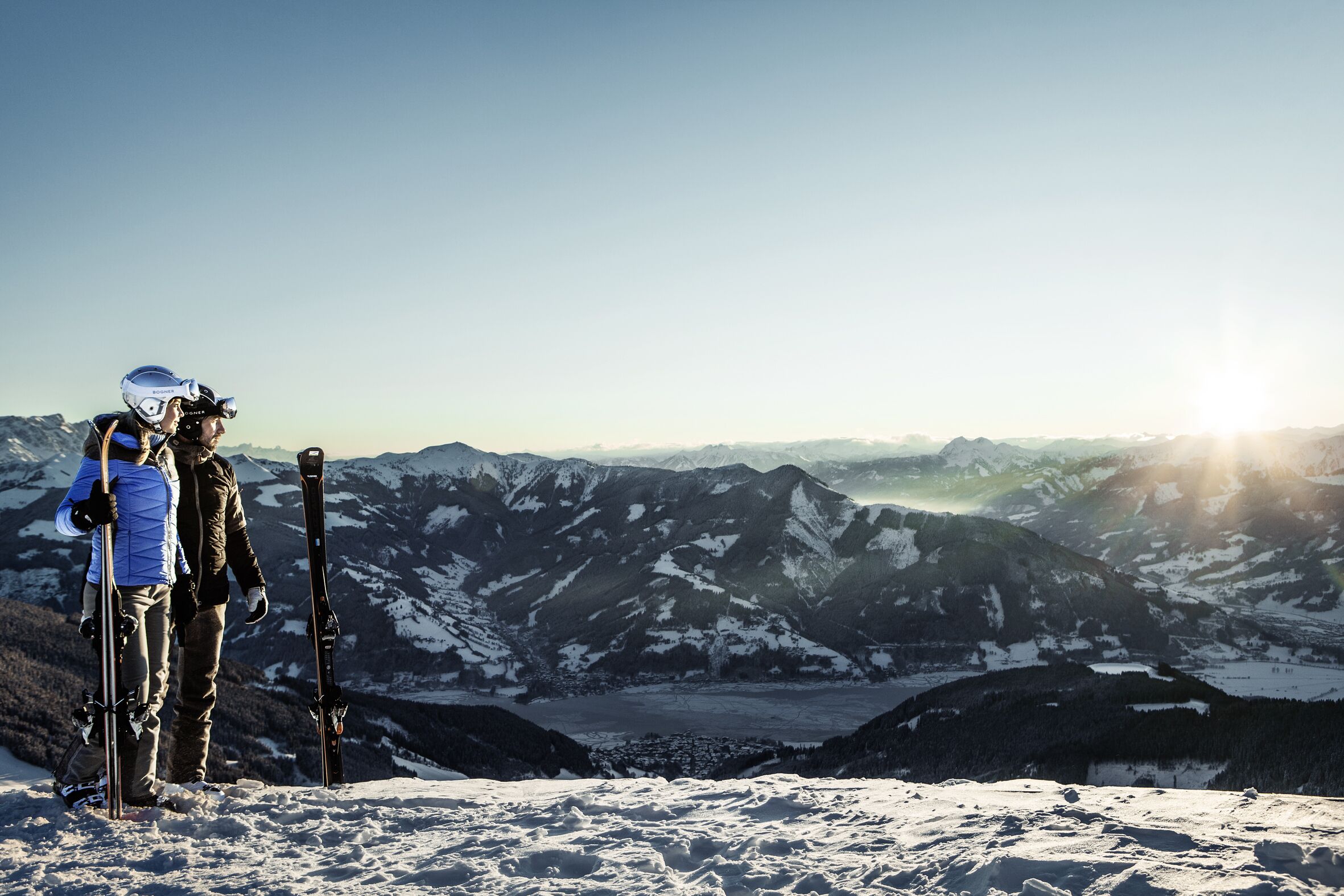 A snowy mountain landscape with two skiers standing on a peak, overlooking a valley with a glacier.