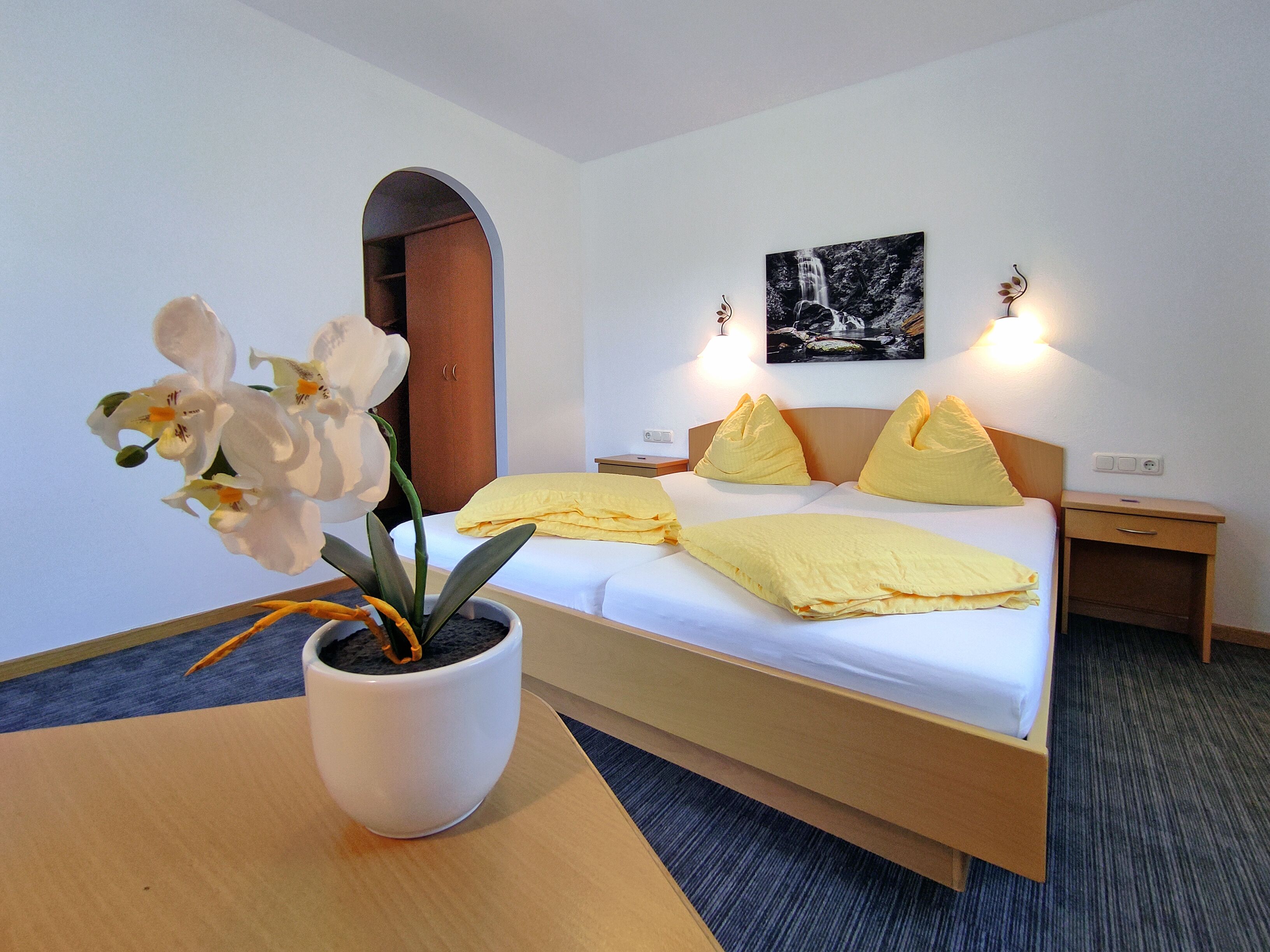 Habitación de hotel con cama, mesita de noche con una orquídea en maceta, lámpara de pared e imagen enmarcada en la pared.