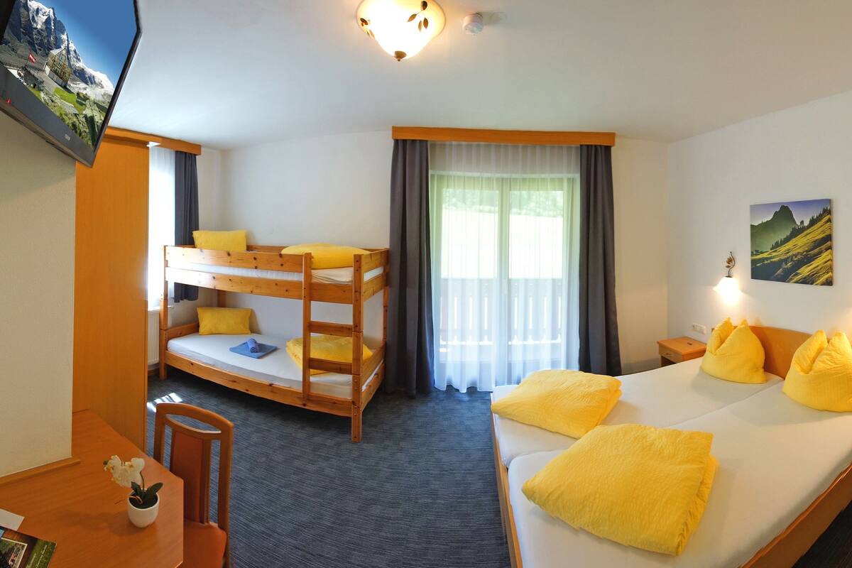 A hotel room with a bunk bed, a single bed, a TV, a desk with a chair, and a window with curtains.