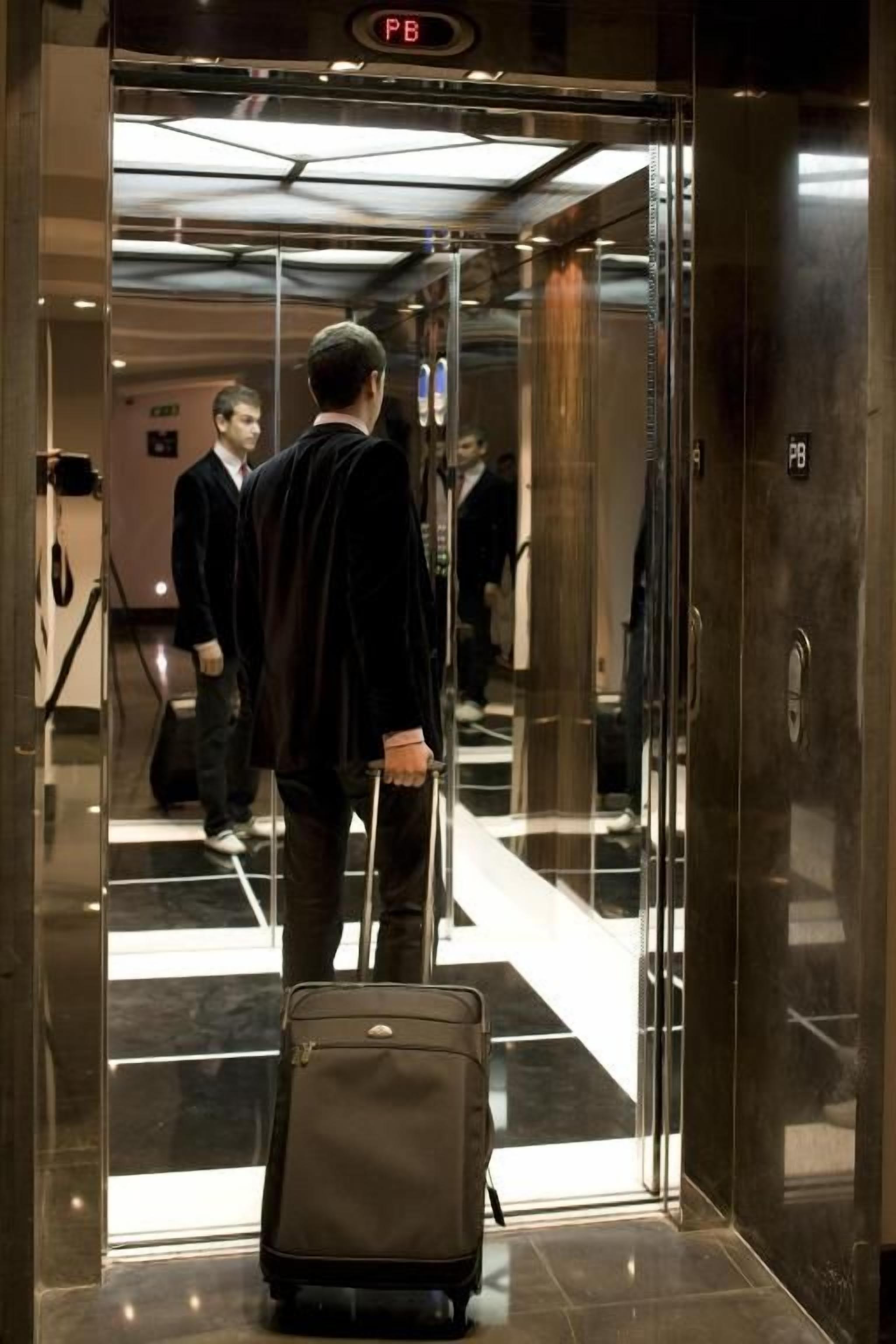 A man in a black suit with a briefcase entering a glass elevator.