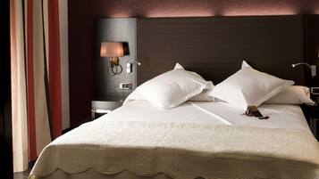Una cama bien hecha con ropa blanca y un cabecero decorativo. Se observa una mesita de noche con una lámpara y un pequeño adorno sobre la cama.