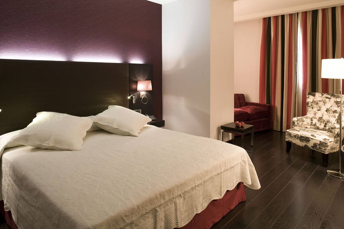 Una habitación de hotel con una cama grande, una mesita de noche con lámpara, un sillón rojo y un sillón estampado.
