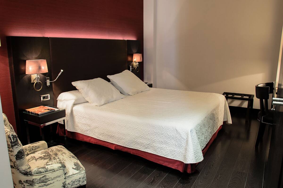 Una cama bien tendida con colcha blanca, dos mesitas de noche con lámparas, una silla con estampado floral y un piso de madera oscura.