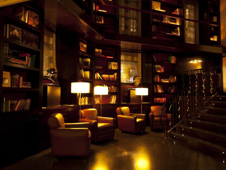 A dimly lit library with leather chairs, bookshelves, and a staircase.
