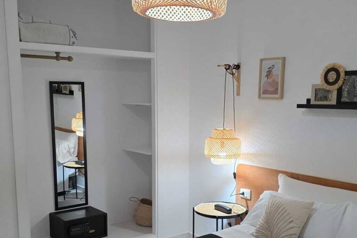 Un dormitorio con una cama negra, un espejo grande, una mesita de noche con lámpara y un aplique colgante.