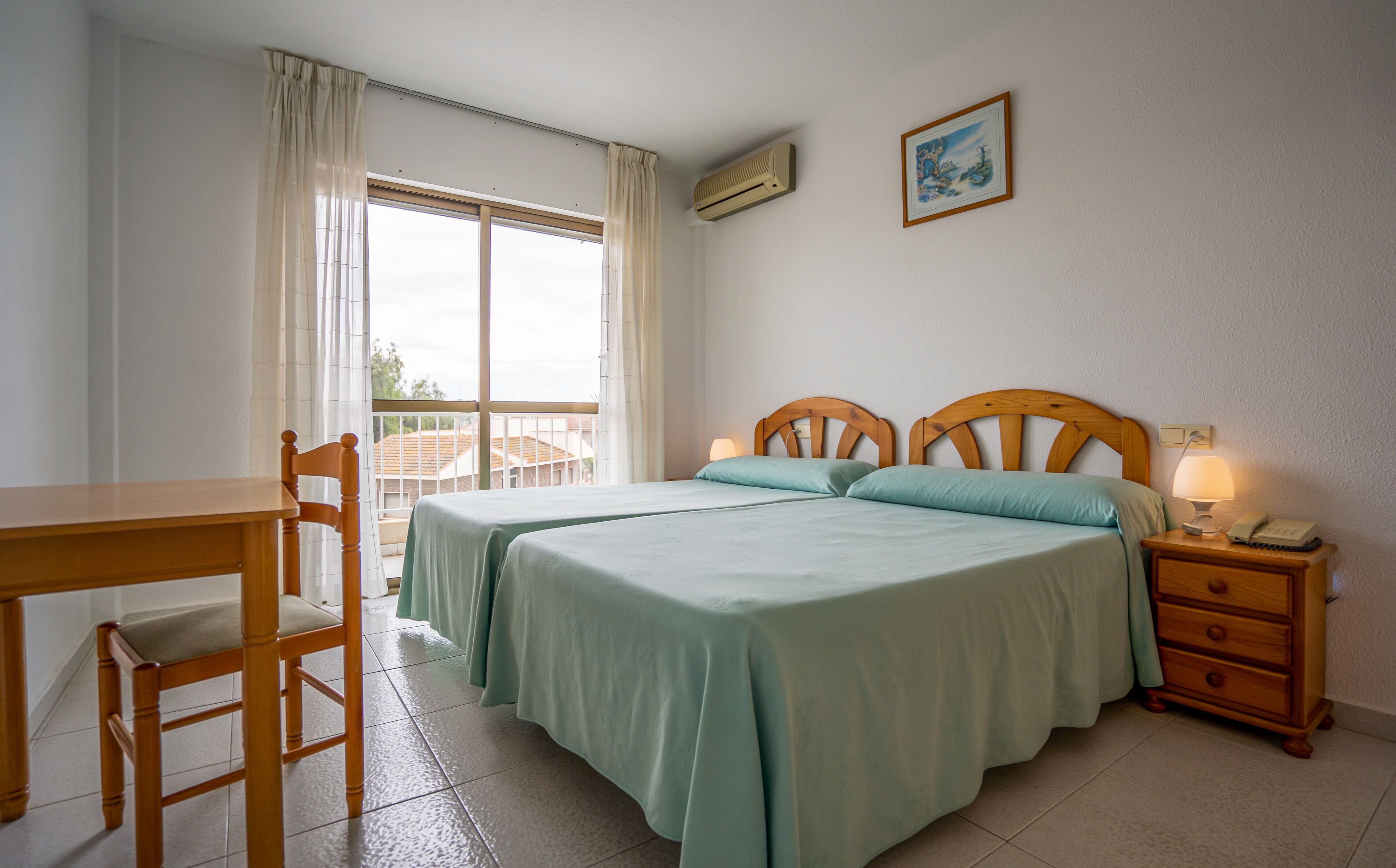 A bedroom with two beds, a desk, a chair, a bedside table with a lamp, and a picture on the wall.