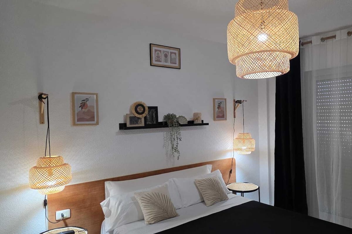 Un dormitorio con una cama, dos lámparas de noche, una mesita de noche, una estantería con objetos decorativos y una ventana con cortinas.