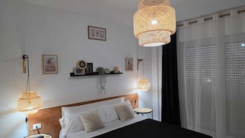 Un dormitorio con una cama, dos lámparas de noche, una mesita de noche, una estantería con objetos decorativos y una ventana con cortinas.
