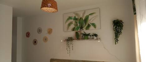 A room with a woven pendant light, a framed painting of tropical leaves, a small shelf with plants, and decorative wall art.