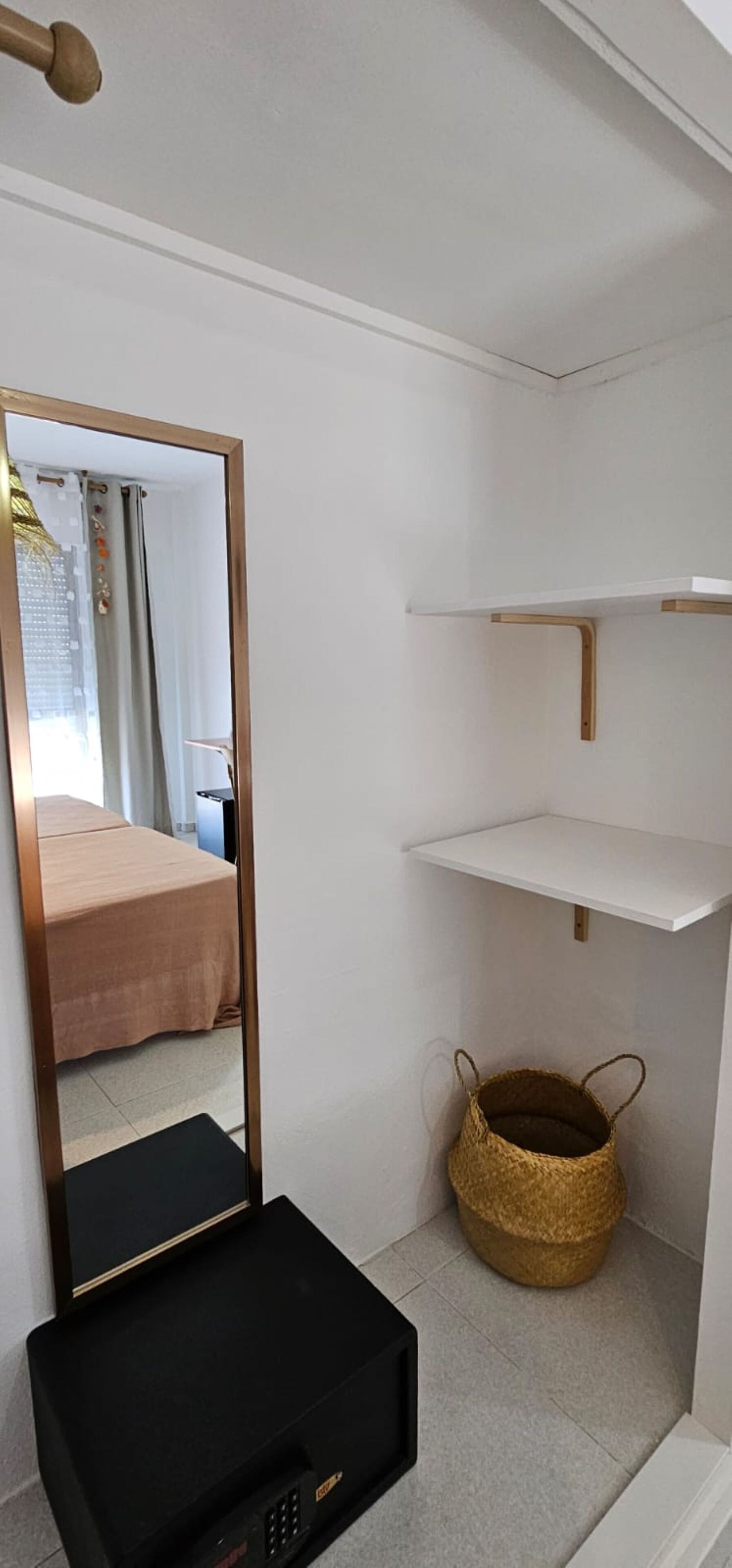 Een kleine kamer met een bed, een spiegel, een klein zwart elektronisch apparaat, een geweven mand en witte planken.