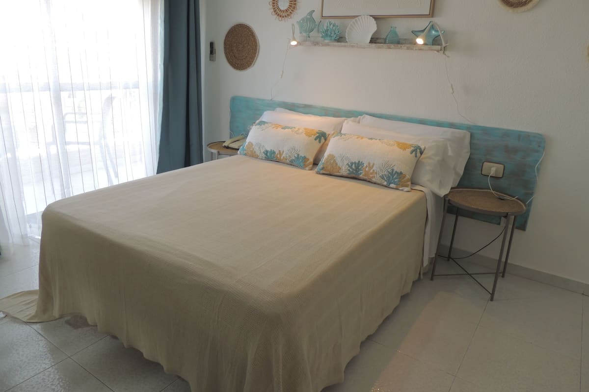 Un dormitorio con una cama grande, dos mesitas de noche, una mesita auxiliar, un estante montado en la pared con adornos y una ventana con cortinas.