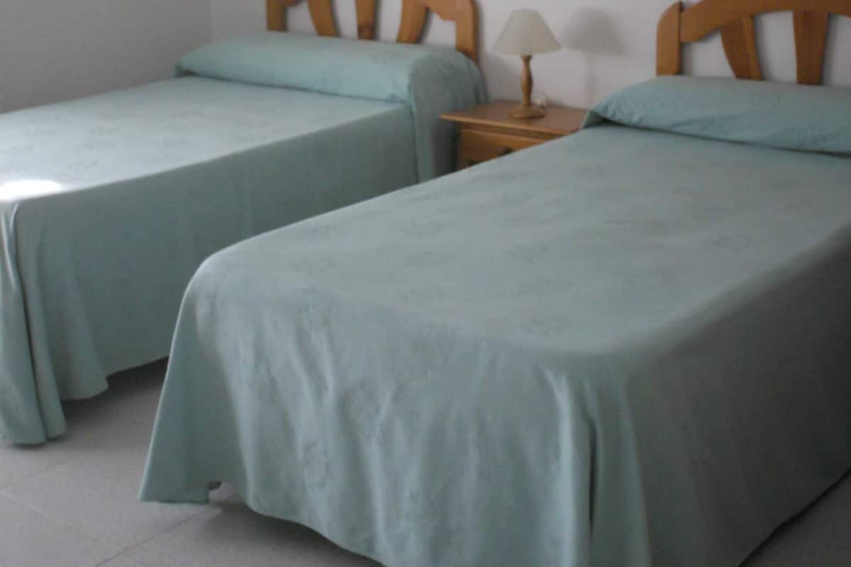 Una habitación con dos camas individuales, cada una con cabecero de madera y una colcha azul claro. Entre las camas, una mesita de noche de madera con una lámpara.
