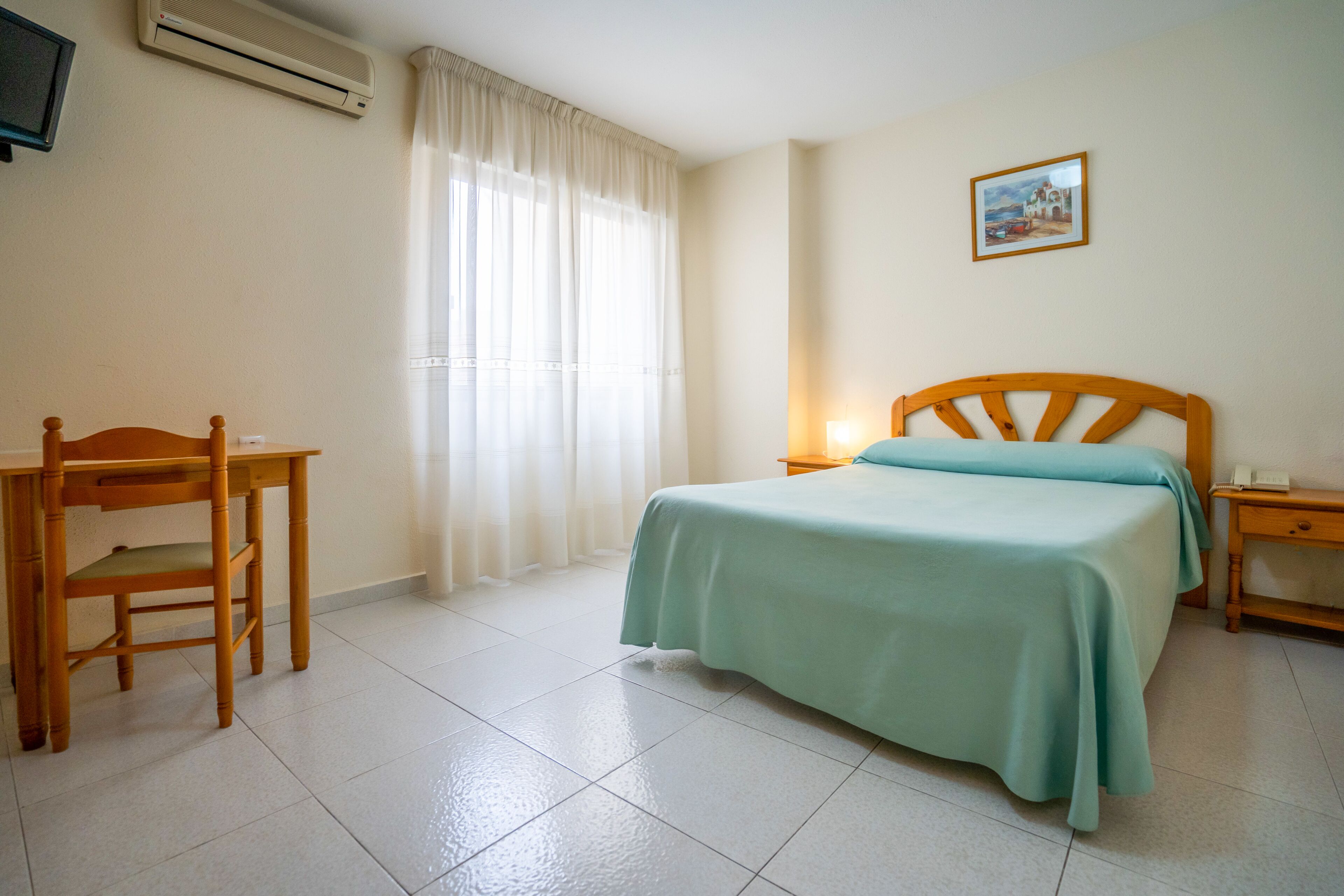 Una habitación de hotel con cama, escritorio, silla, mesita de noche y televisor montado en la pared.