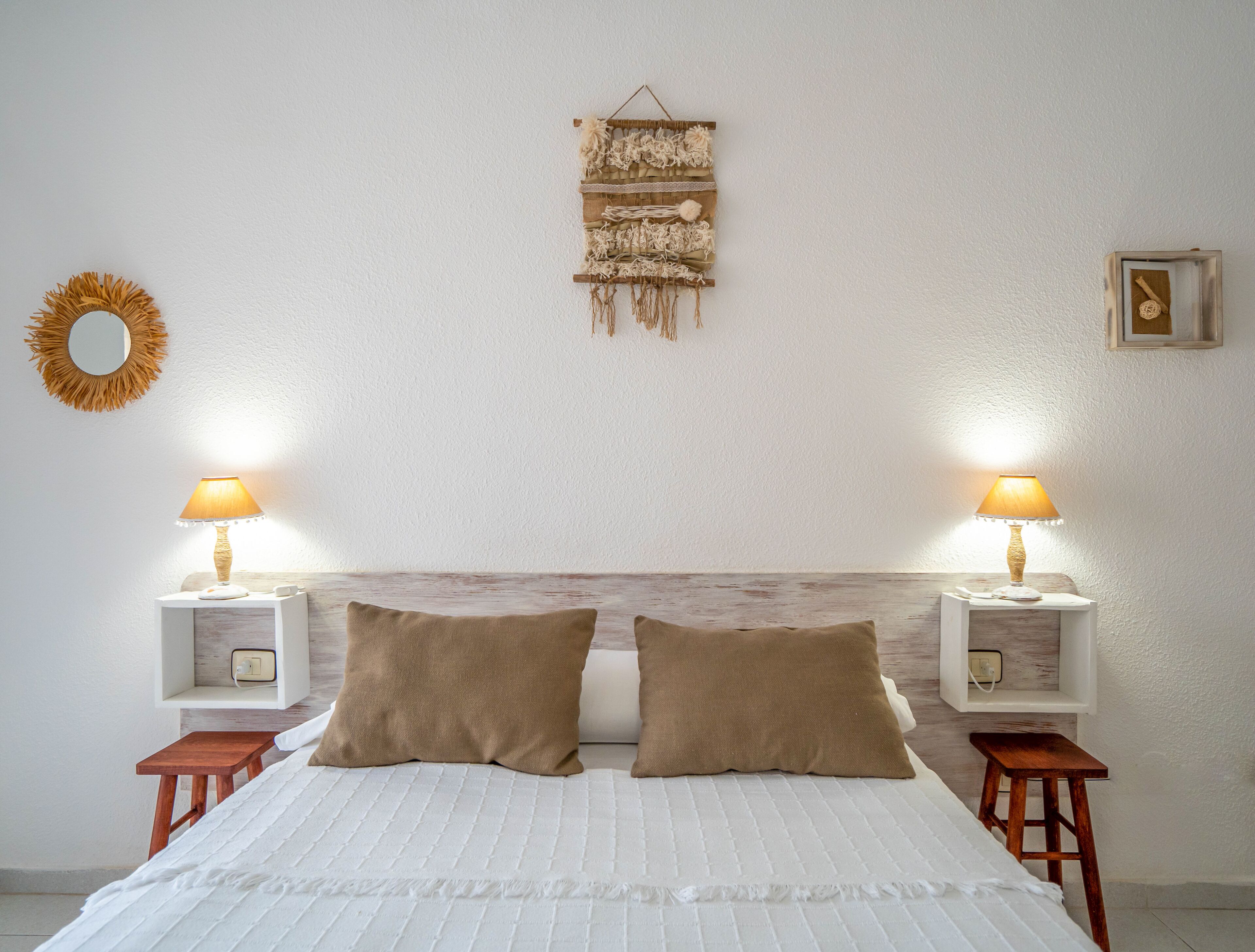 Quarto com cama, duas mesinhas de cabeceira, duas luminárias, dois travesseiros, um painel decorativo na parede e um espelho.